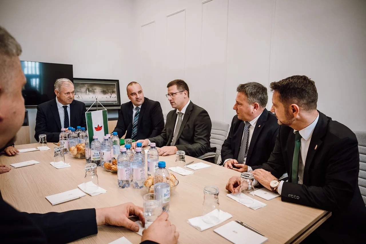 Öltönyös férfiak ülnek egy tárgyalóasztal körül egy irodai teremben, tárgyalás közben. Az asztalon ásványvizes üvegek, kávéscsészék, szalvéták és egy kis székelyzászló látható.