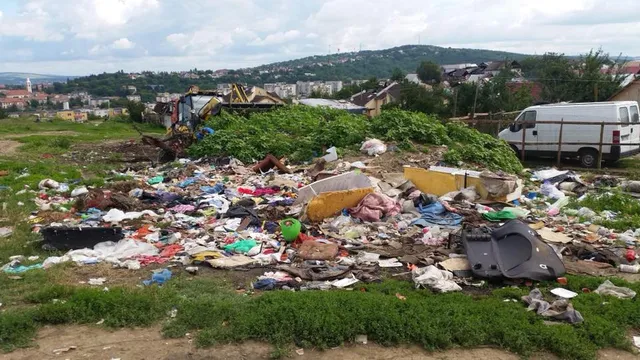 Illegális szemétlerakó egy zöld domboldalon, szanaszét szórt hulladékkal, háttérben egy kotrógép és egy fehér kisteherautó, a távolban Marosvásárhely városképe és dombok láthatók.