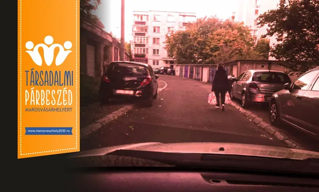 Autó szélvédőjén keresztül fotózott utcarészlet lakóépületek és garázsok között; parkoló autók sorakoznak az úttest szélén, egy bevásárlószatyrokkal sétáló nő halad háttal a kamerának.