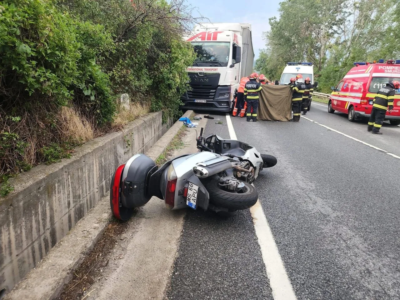 Közúti baleset helyszíne: az úttest szélén felborult, súlyosan sérült motorkerékpár hever, mögötte tűzoltók és mentősök egy takaróval eltakart tárgy körül állnak, a háttérben kamion, mentőautó és tűzoltóautó parkol az út szélén.