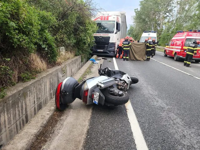 Közúti baleset helyszíne: az úttest szélén felborult, súlyosan sérült motorkerékpár hever, mögötte tűzoltók és mentősök egy takaróval eltakart tárgy körül állnak, a háttérben kamion, mentőautó és tűzoltóautó parkol az út szélén.