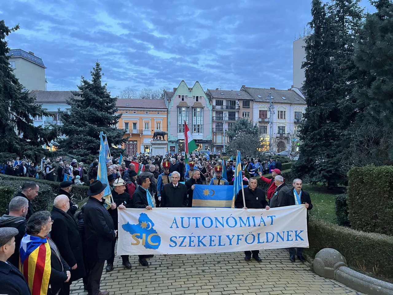 Nagy létszámú tömeg vonul egy téren esti szürkületben, elöl férfiak székely zászlót és „AUTONÓMIÁT SZÉKELYFÖLDNEK” feliratú transzparenst tartanak, a háttérben történelmi épületek és a farkasos-oszlopos szobor látszik.