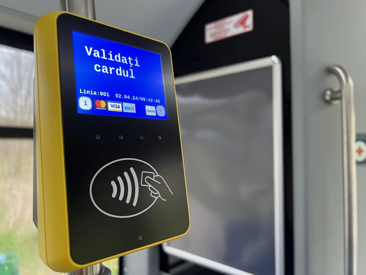 Közeli kép egy tömegközlekedési buszban elhelyezett bankkártya-validáló készülékről, amelynek kijelzőjén román nyelvű felirat olvasható: "Validați cardul" (Validálja a kártyát), valamint a vonal száma (001) és a dátum, idő (02.04.24/09:42:48). A készülék alján egy érintés nélküli fizetést jelző szimbólum és egy kéz sematikus rajza látható.