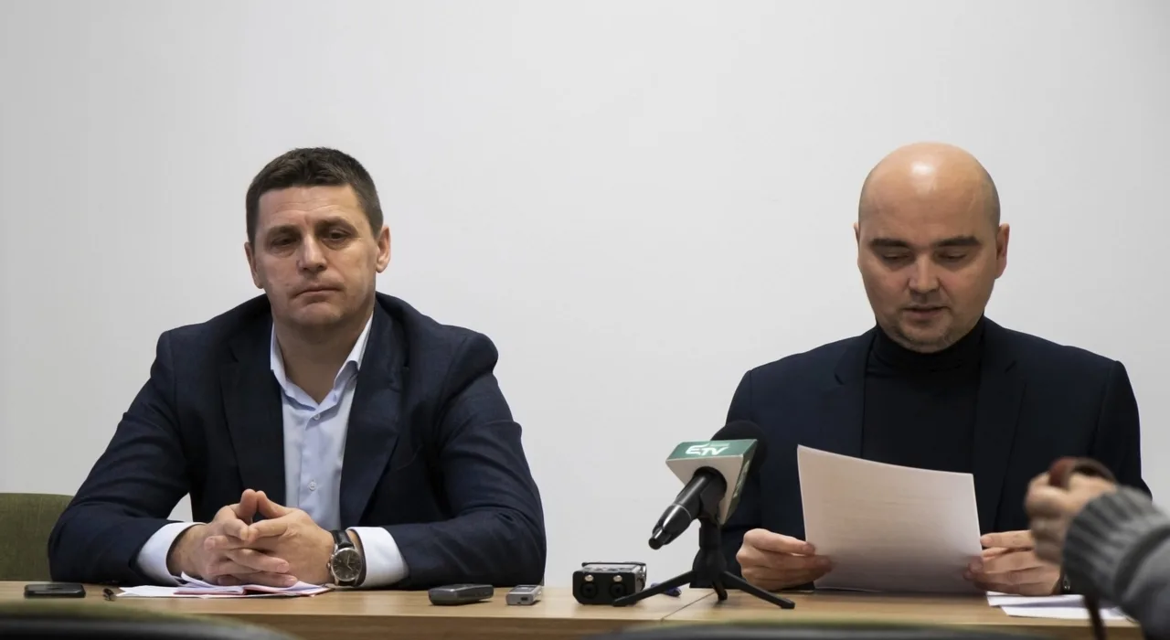 Két öltönyös férfi ül egy asztalnál sajtótájékoztatón, előttük mikrofonok és diktafonok. A jobb oldali férfi papírlapról olvas, a bal oldali kezeit összekulcsolva néz maga elé.