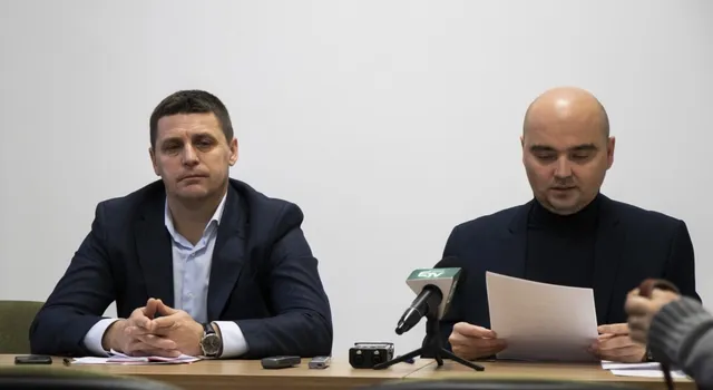 Két öltönyös férfi ül egy asztalnál sajtótájékoztatón, előttük mikrofonok és diktafonok. A jobb oldali férfi papírlapról olvas, a bal oldali kezeit összekulcsolva néz maga elé.