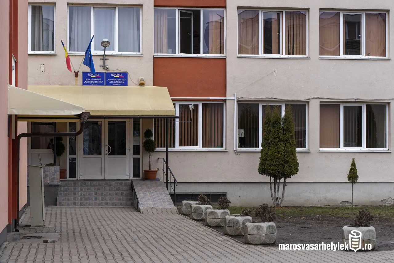 Az Alexandru Ioan Cuza Általános Iskola bejárata Marosvásárhelyen, bézs színű ernyővel fedett bejárati lépcsővel, rámpával, kétnyelvű (román-magyar) feliratú táblákkal az ajtó felett, valamint román és uniós zászlóval.