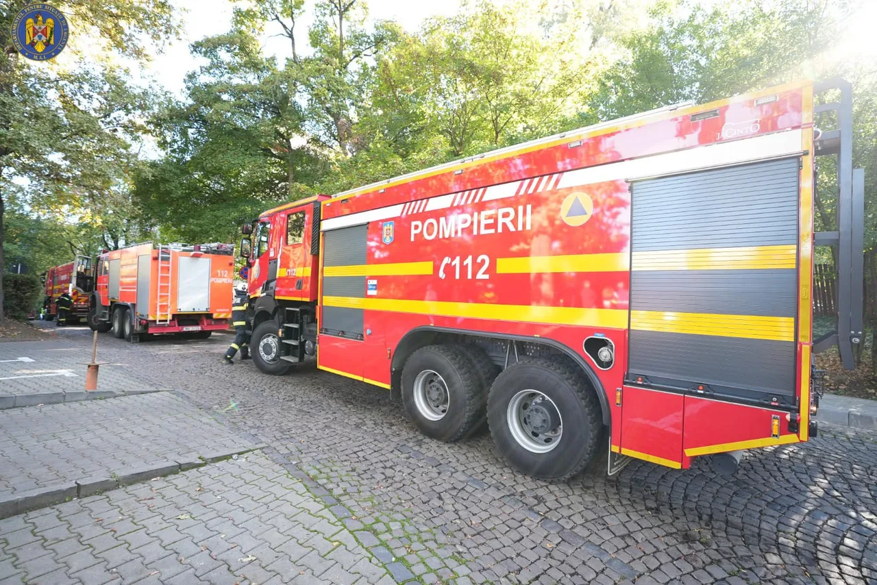 Nagy méretű piros-sárga tűzoltóautók (Pompierii 112 felirattal) parkolnak egy kockakővel fedett úton, fák alatt, a háttérben egy másik tűzoltóautó és egy tűzoltó látható.