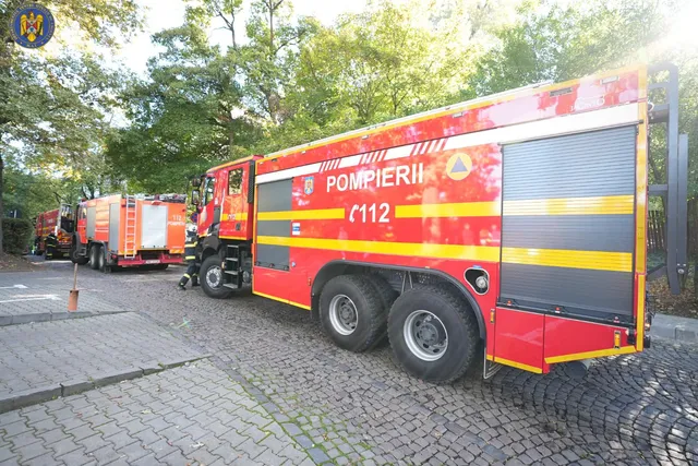 Nagy méretű piros-sárga tűzoltóautók (Pompierii 112 felirattal) parkolnak egy kockakővel fedett úton, fák alatt, a háttérben egy másik tűzoltóautó és egy tűzoltó látható.