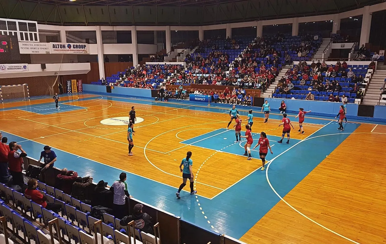 Sportcsarnokban zajló női kézilabdamérkőzés felülnézetből, a pályán piros és kék-fekete mezes játékosok, a lelátókon nézők ülnek.
