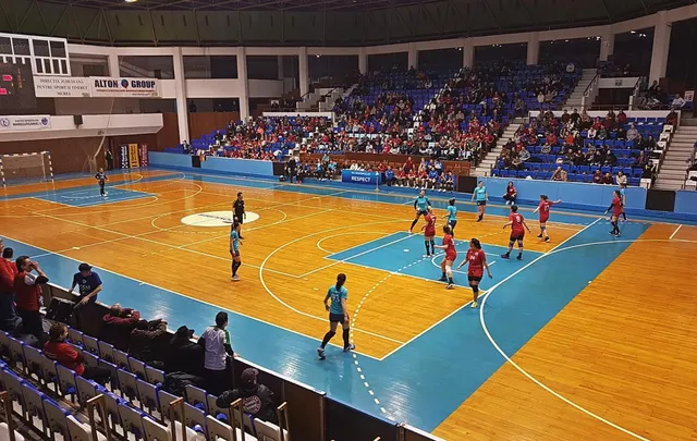 Sportcsarnokban zajló női kézilabdamérkőzés felülnézetből, a pályán piros és kék-fekete mezes játékosok, a lelátókon nézők ülnek.