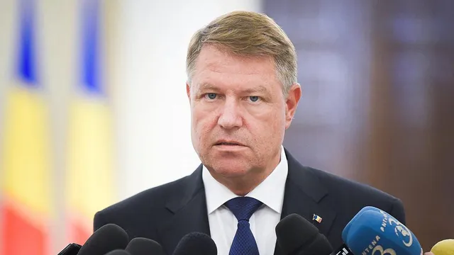 Öltönyt és nyakkendőt viselő férfi mikrofonok előtt beszél, mögötte román zászlók láthatók.