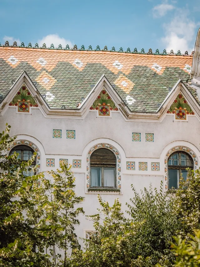 Szecessziós stílusú épület homlokzatának részlete, három csúcsíves ablakkal és háromszög alakú oromzattal, mázas cserépből kirakott mintás tetővel, díszes csempeornamentikával. Az előtérben zöld lombozat takarja az alsó részt.
