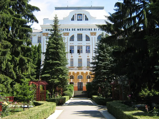 Fehér, klasszicista stílusú épület homlokzata, rajta az „UNIVERSITATEA DE MEDICINĂ ȘI F...” felirat, előtte ápolt park, nyírott sövények, fenyőfák és egy sétány vezet a bejárathoz.