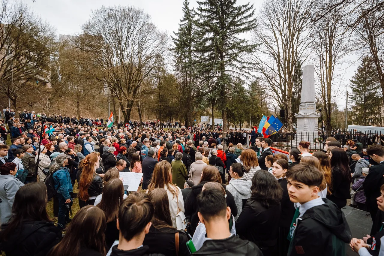 Nagy tömeg gyűlik össze egy parkban a Székely vértanúk emlékművénél, március 15-i megemlékezésen, magyar és uniós zászlókkal, kopár fákkal a háttérben.