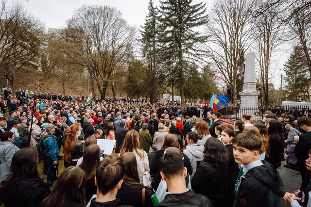 Nagy tömeg gyűlik össze egy parkban a Székely vértanúk emlékművénél, március 15-i megemlékezésen, magyar és uniós zászlókkal, kopár fákkal a háttérben.