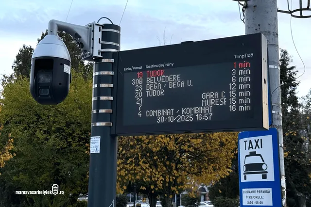 Buszmegálló digitális kijelzője látható egy oszlopon, amely a járatok számát, célállomását és érkezési idejét mutatja; mellette biztonsági kamera és taxiállomás jelzőtábla van elhelyezve, háttérben őszi lombozatú fák.