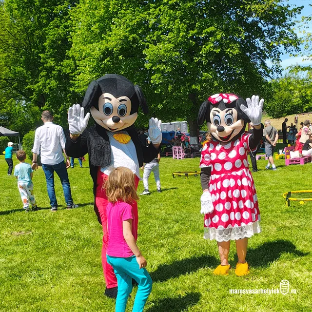 Egyértelműen felismerhető Mickey egér és Minnie egér jelmezes maskotta integet egy zöld parkban, előttük egy kislány áll, háttérben emberek és rendezvénysátrak láthatók napsütéses időben.