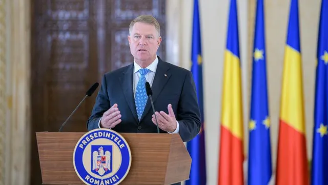 Öltönyt és nyakkendőt viselő férfi beszél egy pulpituson, amelyen 'Președintele României' felirat és címer látható, mögötte román és európai uniós zászlók.