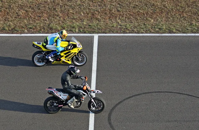 Felülnézeti kép egy versenypályáról, amelyen két motorkerékpáros halad: elöl egy fekete-fehér motoron ülő versenyző fekete overallban, mögötte egy sárga sportmotoron ülő versenyző fehér-kék-sárga overallban és sisakban.