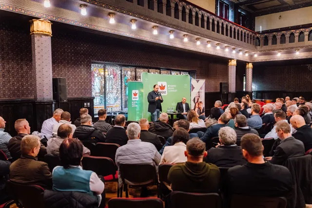 Zsúfolt, díszes teremben (üvegfestett ablakokkal, mór stílusú karzattal) egy férfi mikrofonnal beszél az RMDSZ Maros megyei szervezetének zöld logós háttere előtt, mellette egy másik férfi ül egy kisasztalnál; előttük nagyszámú közönség ül és hallgatja.