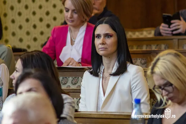 Ülésteremben, fapados padsorokban ülő nők egy csoportja, előtérben egy fekete hajú nő fehér blézerben figyel komoly arckifejezéssel, mögötte egy pink blézeres nő jegyzeteket néz.