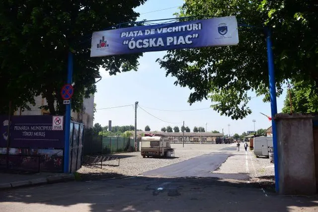 Bejárat kapuja a marosvásárhelyi ócskapiacnak, felette kék táblán „Piata de vechituri – Ócska piac” felirat, mögötte kavicsos térség, lakókocsi és néhány épület.