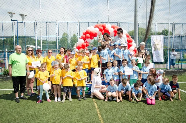 Nagy csoport óvodás és iskolás gyermek áll és guggol csoportképen egy műfüves sportpályán, sárga és világoskék pólókban, néhányan okleveleket tartanak; mögöttük léggömbökből készült piros-fehér ív és felnőttek láthatók.
