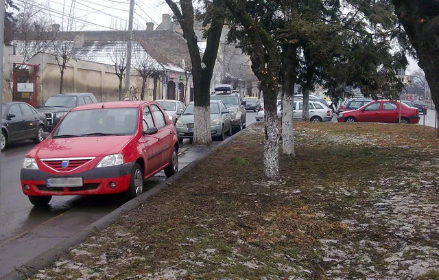 Utcán, füves-fás terület mellett parkoló autók sorakoznak a járda mentén, előtérben egy piros kisautó, háttérben régi épületek és villanyoszlopok.