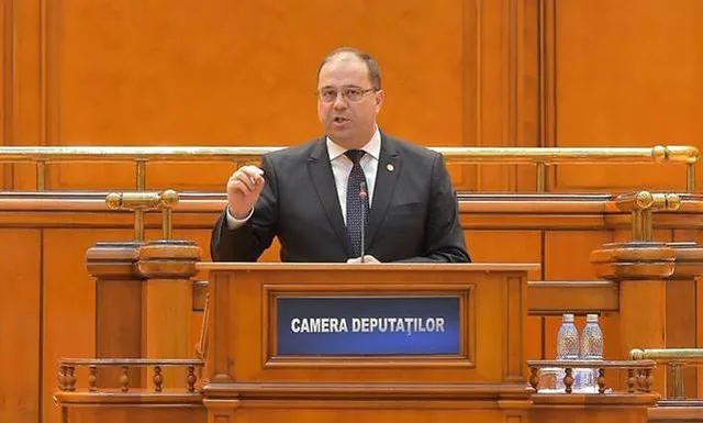 Öltönyt viselő férfi beszél a Camera Deputaților (Képviselőház) felirattal ellátott pulpitusnál a román parlament üléstermében, kezét gesztikulálva emeli.