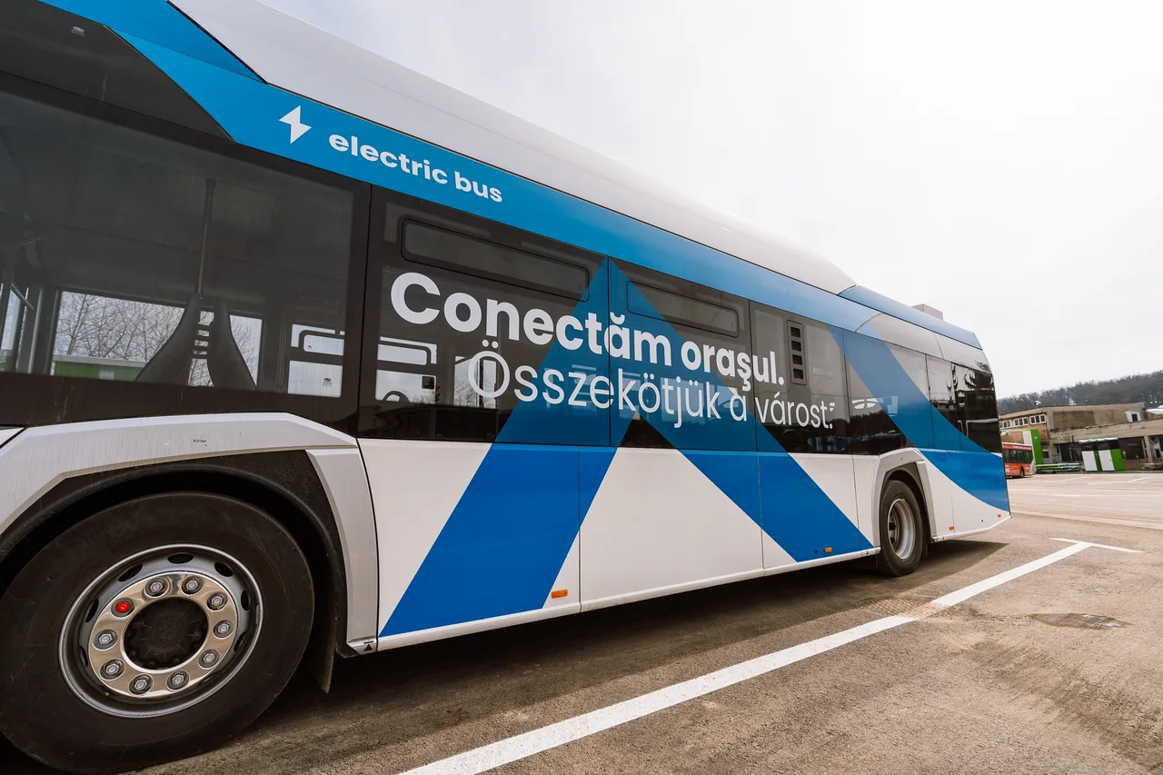 Kék-fehér elektromos autóbusz oldalnézetből, oldalán kétnyelvű felirat: „Conectăm oraşul. Összekötjük a várost.” és „electric bus”, egy parkolóban áll.