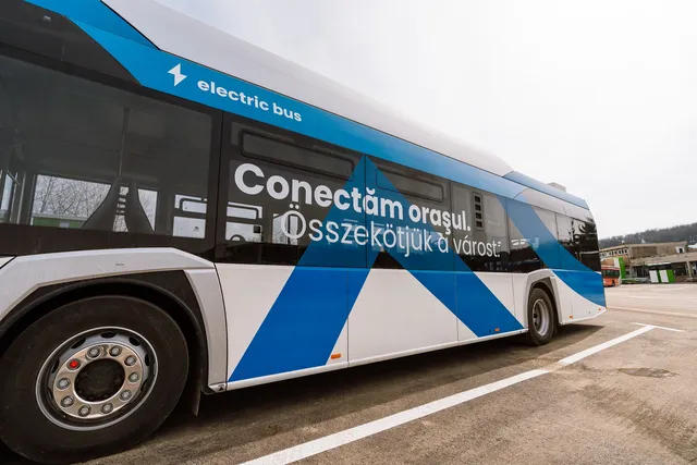 Kék-fehér elektromos autóbusz oldalnézetből, oldalán kétnyelvű felirat: „Conectăm oraşul. Összekötjük a várost.” és „electric bus”, egy parkolóban áll.