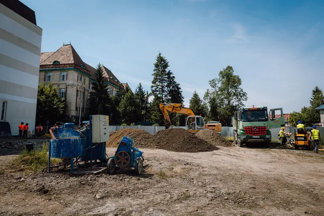 Építkezési terület, kotógép és két teherautó, homokkupacok, betonkeverő gép, munkások láthatók a háttérben egy régi épület és fenyőfák előtt.