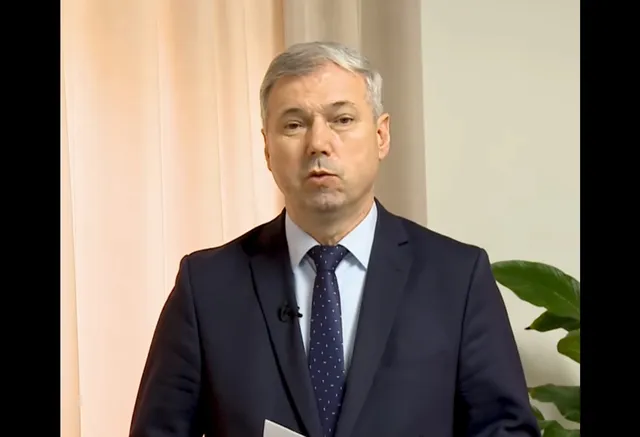 Sötét öltönyt és mintás nyakkendőt viselő középkorú férfi beszél kamera előtt, mögötte bézs függöny és egy zöld növény látható.