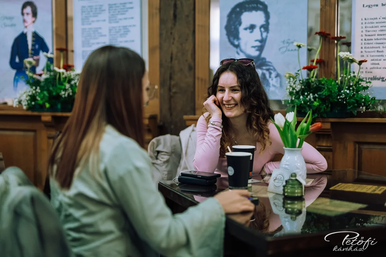 Két nő beszélget egy kávéház asztalánál, kávés poharakkal és egy vázába helyezett tulipánokkal az asztalon, a háttérben történelmi portrék és feliratos táblák láthatók a falon.