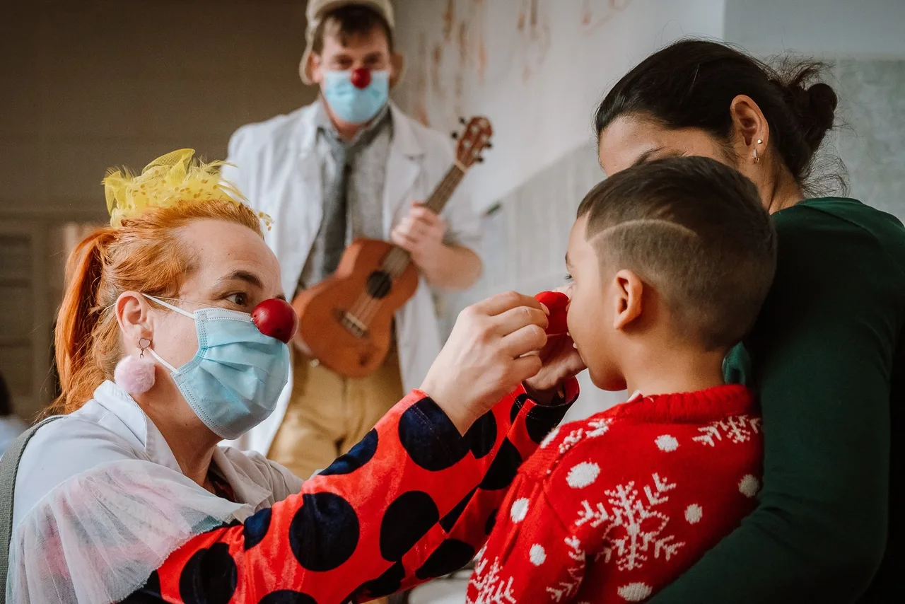 Piros orrú bohócdoktor nő orvosi maszkban piros bohóc orrot tesz fel egy kisfiúnak, mögötte egy másik bohócdoktor ukulelén játszik, a gyermek édesanyja mellette áll.