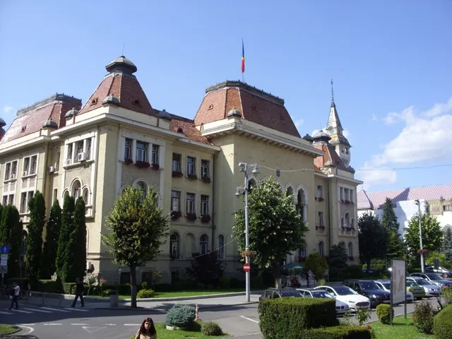 Marosvásárhely régi városháza épülete, jellegzetes tornyokkal és cseréptetőkkel, magyar és román zászlóval a tetőn, előtte parkoló autók és néhány gyalogos.