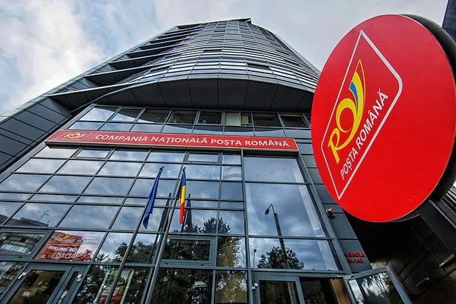 A Román Posta (Poșta Română) székházának épülete alulnézetből fotózva, üvegfalú modern toronyépület, a bejárat felett vörös táblán felirat, előtte román és uniós zászlók; jobb oldalt a cég piros kör alakú logója látható.