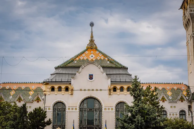 A Kultúrpalota szecessziós stílusú homlokzatának felső része látható, díszes majolika tetőcserepekkel, csúcsán fém dísszel, alatta Románia címere és íves ólomüveg ablakok, előtte fák.