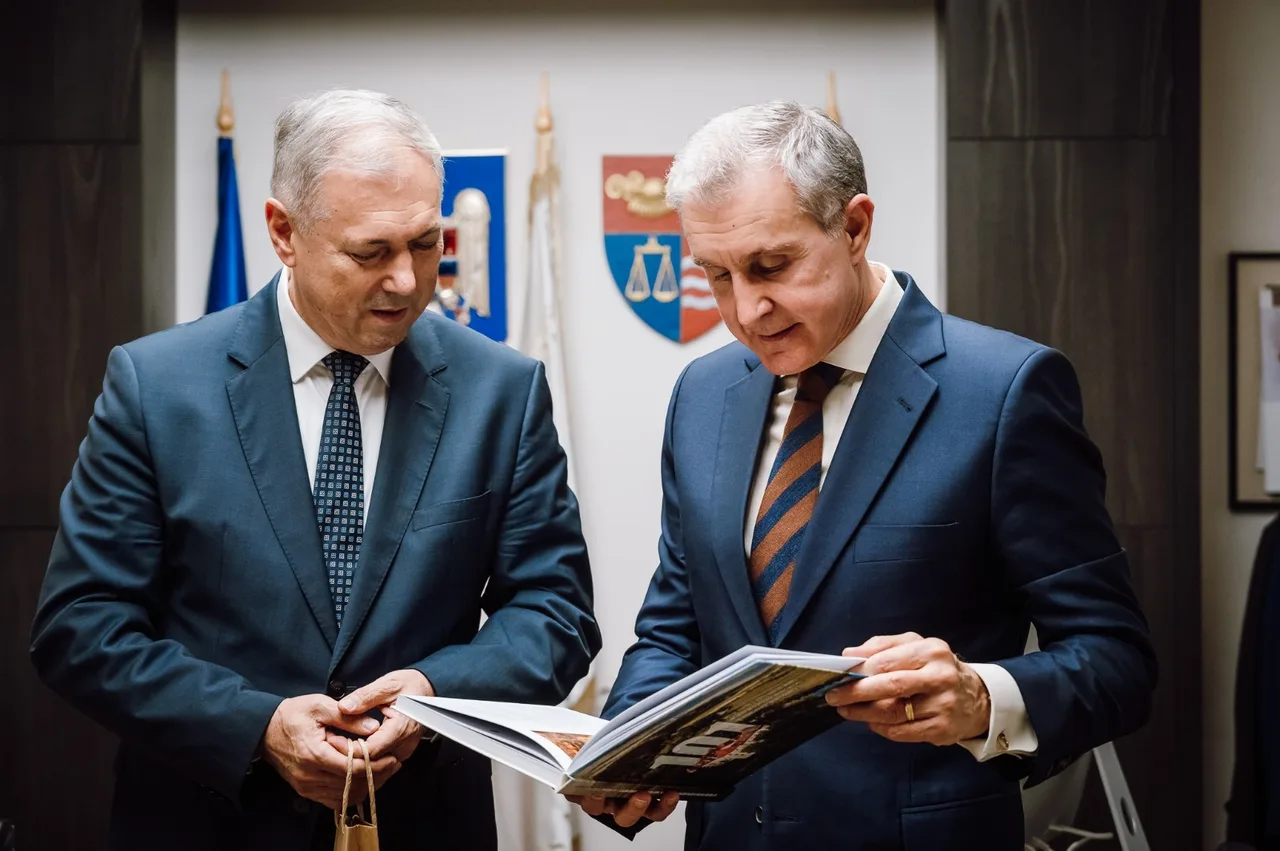 Két öltönyös férfi néz egy nyitott fotóalbumot vagy könyvet, mögöttük Románia zászlaja és Maros megye/Marosvásárhely címere látható egy irodai teremben.