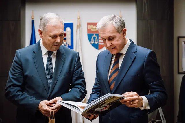 Két öltönyös férfi néz egy nyitott fotóalbumot vagy könyvet, mögöttük Románia zászlaja és Maros megye/Marosvásárhely címere látható egy irodai teremben.