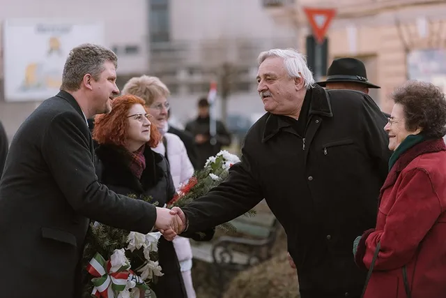 Kültéren emberek kézfogással köszöntik egymást egy megemlékezésen, kezükben nemzeti színű szalagos koszorúk és virágok, háttérben járók és egy régi épület.