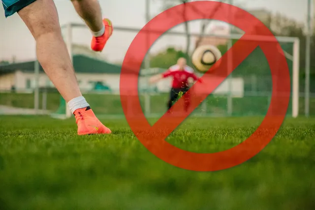Focimeccs jelenete: előtérben egy játékos lába narancssárga stoplis cipőben rúgja a labdát, háttérben egy piros mezes kapus a labda felé vet magát a kapu előtt. A képre nagy piros tiltó jel (áthúzott kör) van montírozva.