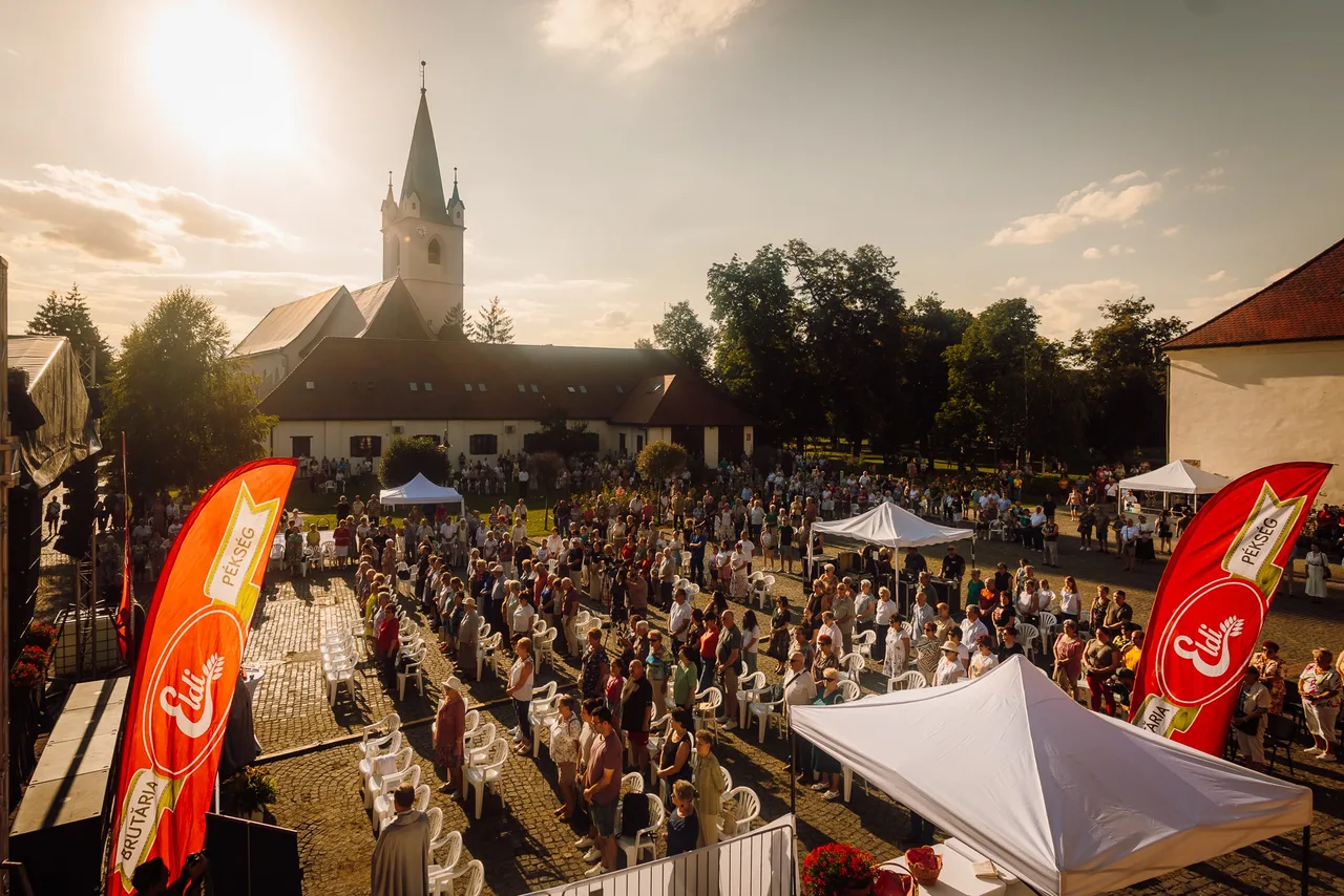 Templom udvarán nagy tömeg gyülekezik szabadtéri rendezvényen, székek sorai és sátrak közt állnak az emberek napnyugtakor, a háttérben a vártemplom tornya látszik, előtérben pékséget reklámozó piros zászlók.
