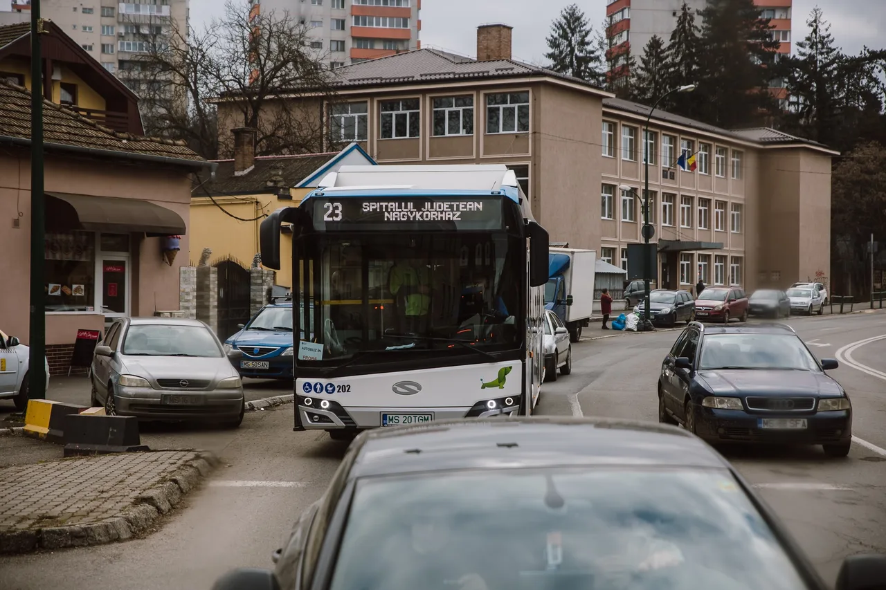 Utcai jelenet Marosvásárhelyen: egy 23-as jelzésű, „Spitalul Judetean / Nagykorház” feliratú városi busz halad az úton, körülötte parkoló és mozgó személygépkocsik, háttérben egy iskola épülete és panelházak.