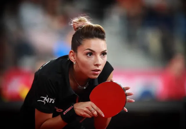 Fiatal nő fekete sportmezben asztaliteniszütőt tart a kezében, koncentráltan figyel előre, háttérben elmosódott nézőtér látszik.
