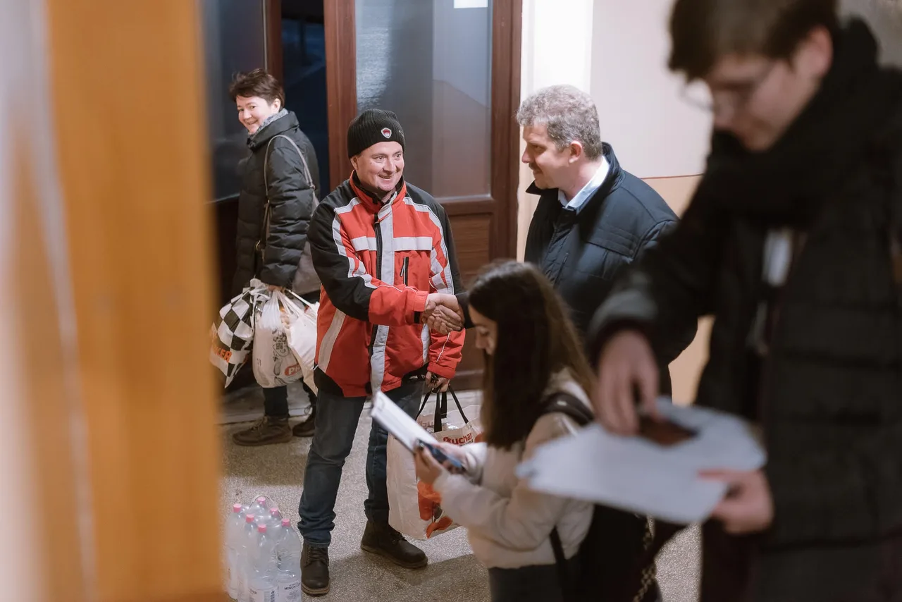 Beltéri helyszínen egy narancssárga-fekete munkaruhás, sapkás férfi kezet fog egy öltönyös, ősz hajú férfival, körülöttük többen bevásárlószatyrokkal és papírokkal állnak, a földön ásványvizes palackok láthatók.