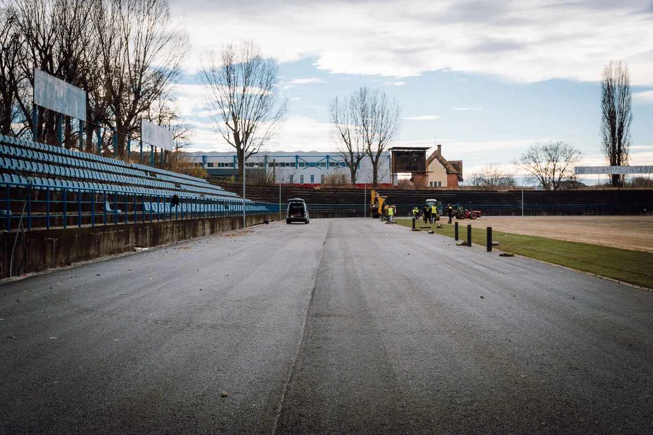 Üres stadion nézőtere kék üléssorokkal, előtte aszfaltozott futópálya, ahol munkások sárga munkaruhában és egy kotrógép dolgozik; háttérben fák és régi épületek láthatók borús égbolt alatt.