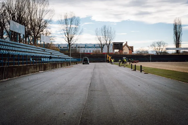 Üres stadion nézőtere kék üléssorokkal, előtte aszfaltozott futópálya, ahol munkások sárga munkaruhában és egy kotrógép dolgozik; háttérben fák és régi épületek láthatók borús égbolt alatt.