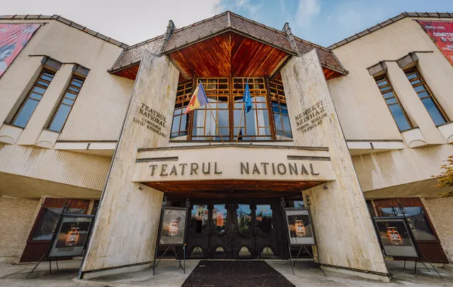 A marosvásárhelyi Nemzeti Színház bejárata, homlokzatán román és magyar nyelvű felirat (Teatrul National / Nemzeti Színház), a bejárat előtt plakátállványok és székely zászló, kék uniós zászló lóg az erkélyen.