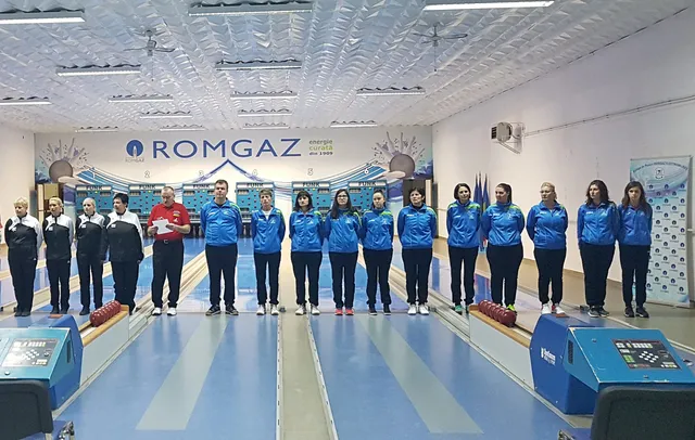 Két csapat tekézője és egy vezető, kék-fehér, illetve kék sportmezben sorakoznak fel egy tekepálya előtt a Romgaz feliratú fal alatt, valószínűleg a mérkőzés előtti köszöntőn.